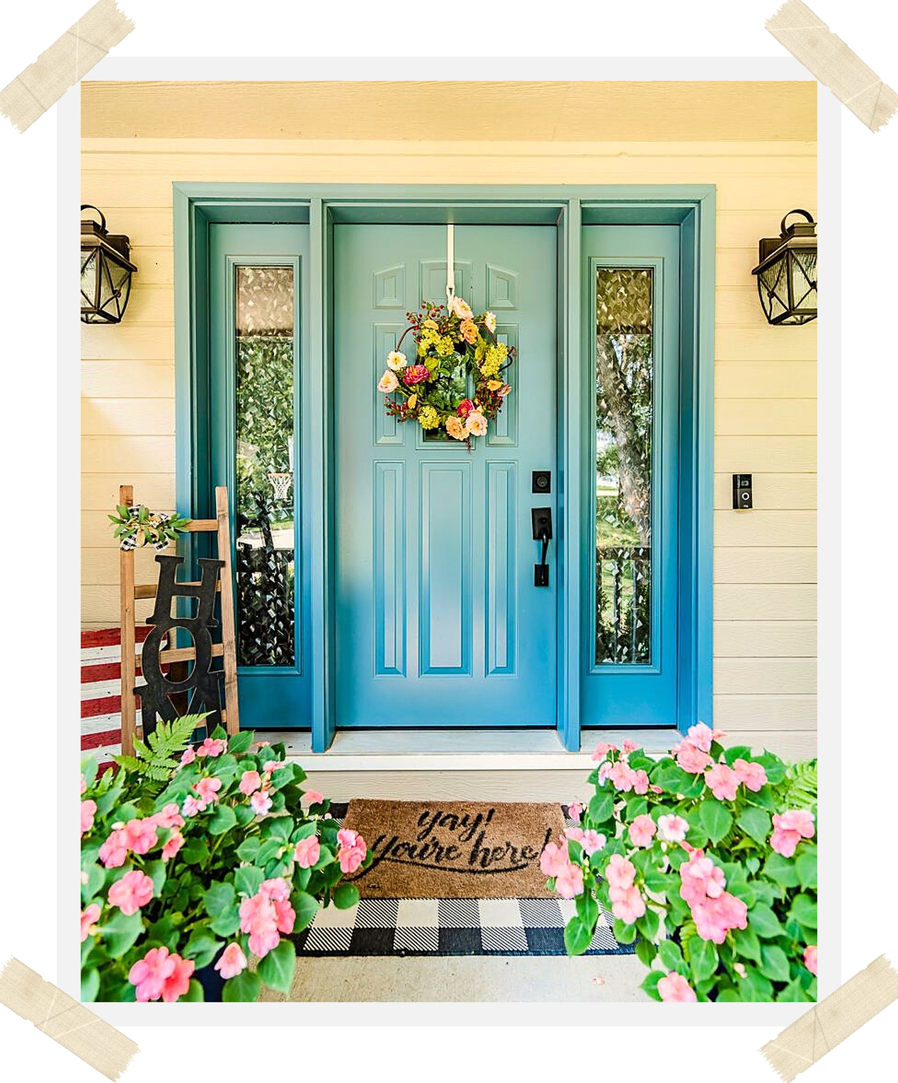 Teal front door with a 'yay! you're here!' doormat and pink flowers on a front porch