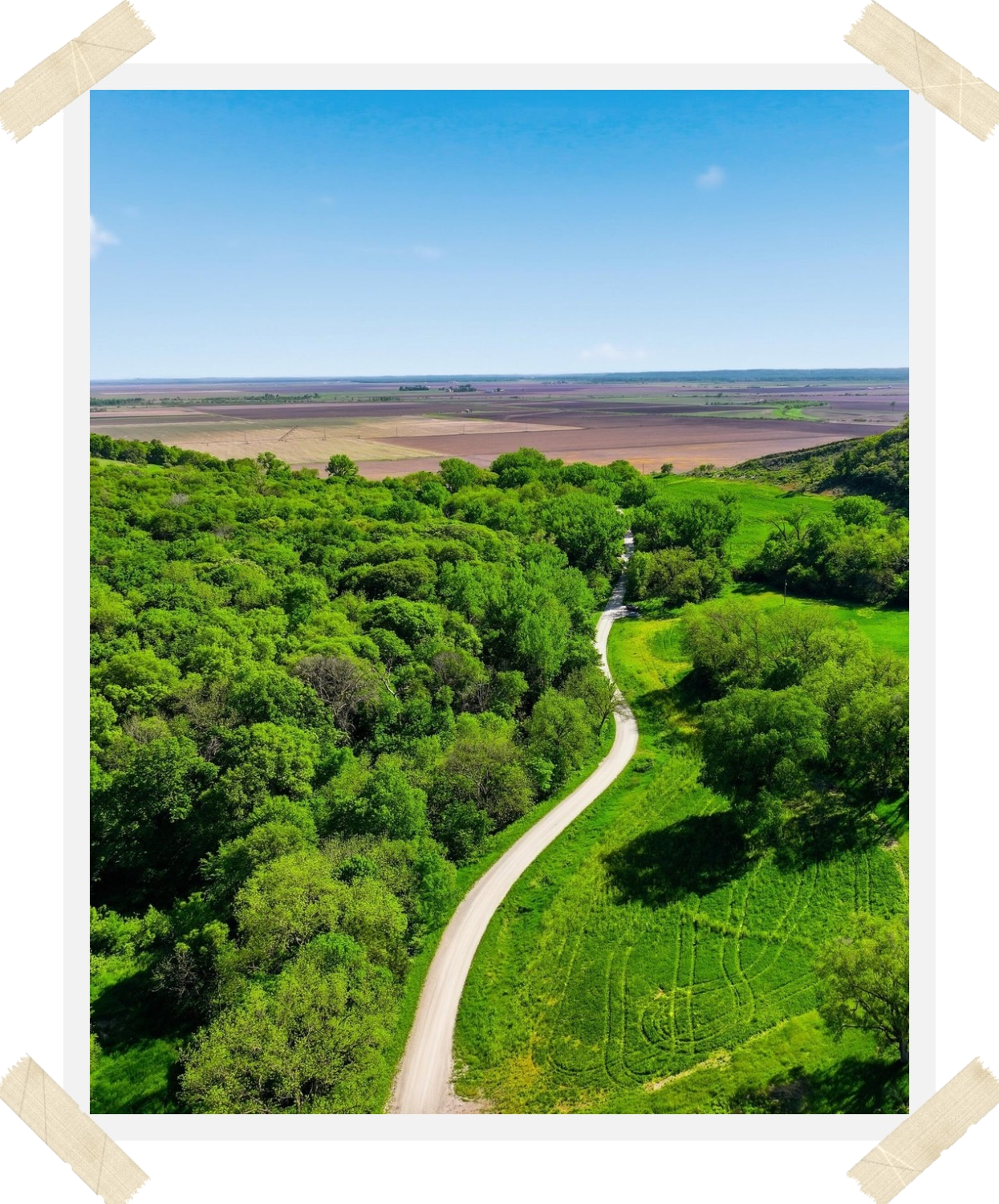 Aerial view of a gravel lane winding through wooded Loess Hills toward open Missouri River bottom farmland in Southwest Iowa